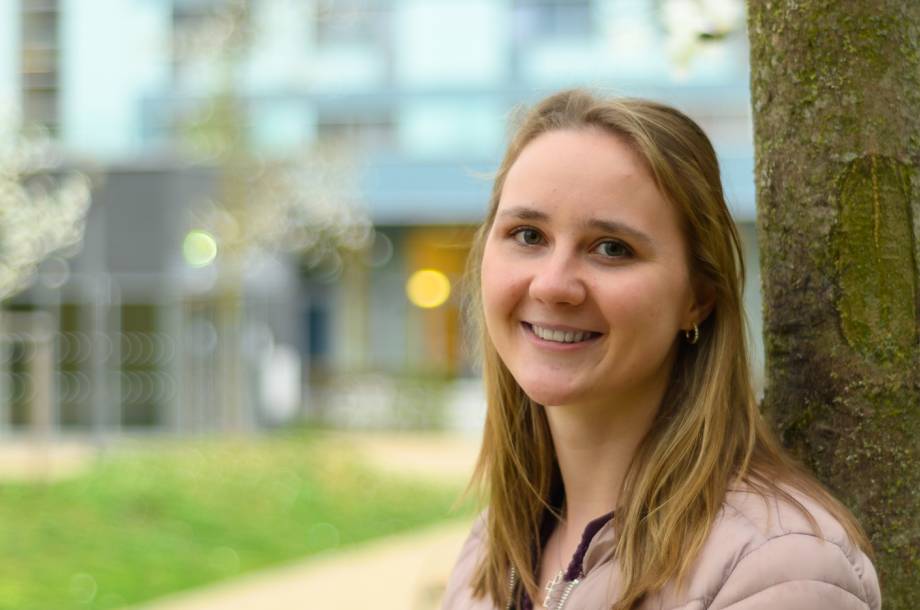 Alexandra lehnt an einem Baum im Park und lächelt in die Kamera.