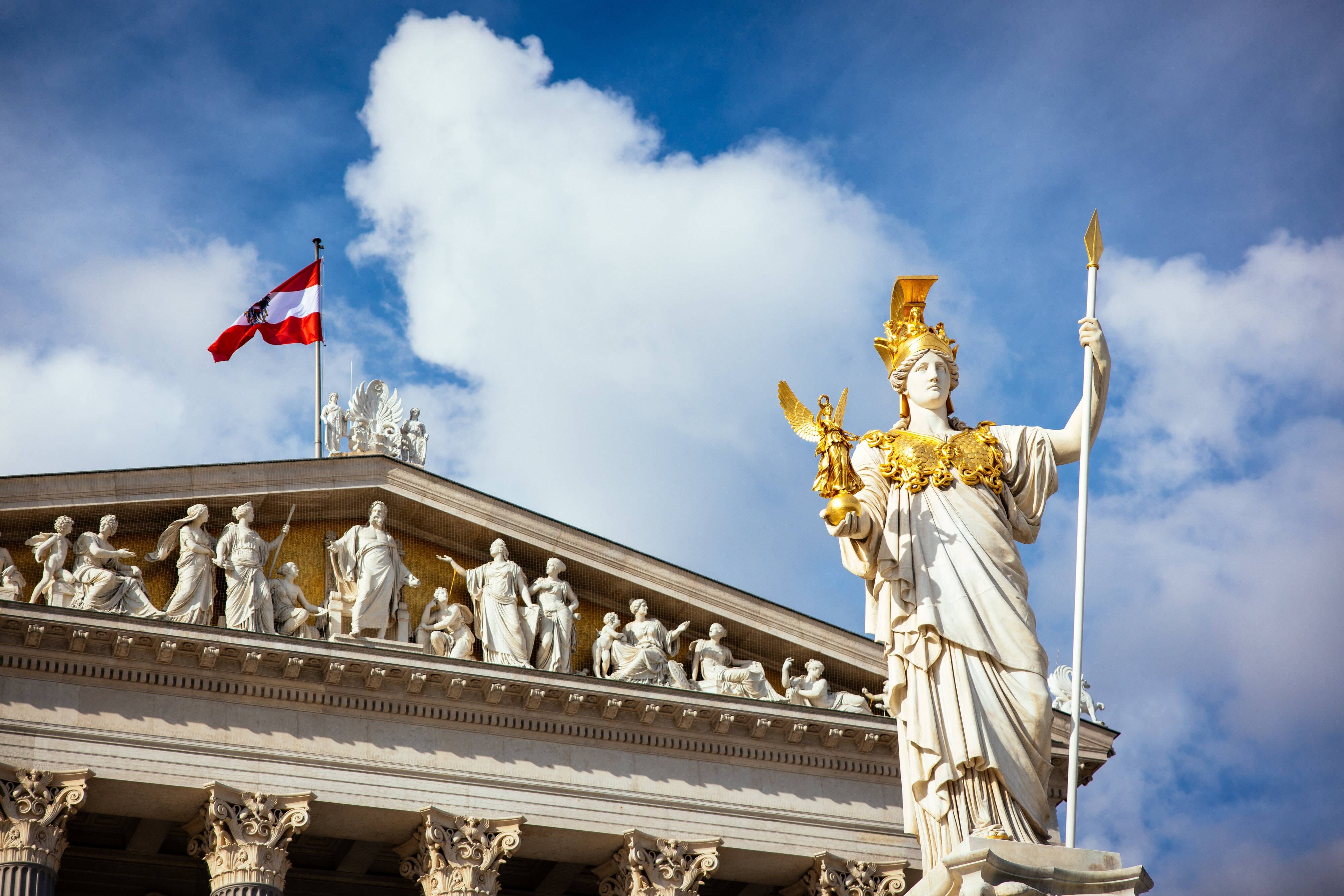 Pallas-Athene-Statue vor dem österreichischen Parlament in Wien mit österreichischer Flagge im Hintergrund unter einem klaren blauen Himmel.